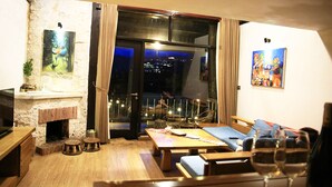 Un salon chaleureux avec une cheminée en pierre, un balcon offrant une vue sur la ville et deux tableaux accrochés aux murs.