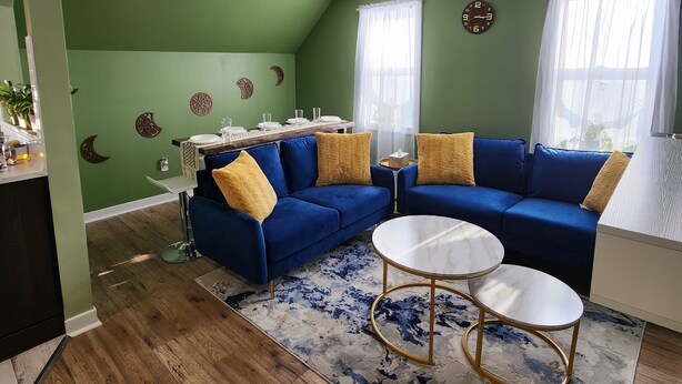 Uma sala de estar com um sofá modular azul, duas mesas de centro redondas, um tapete estampado e um espaço de jantar com uma mesa comprida e cadeiras.