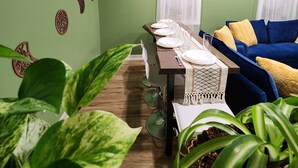 Uma área de refeições com uma mesa comprida posta para quatro pessoas, um sofá azul com almofadas amarelas e várias plantas em vasos.