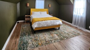 Quarto com cama, mesa de cabeceira, abajur e janela com cortinas translúcidas.