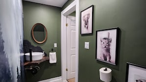 Um banheiro com parede verde, pia branca, espelho e quadros na parede.