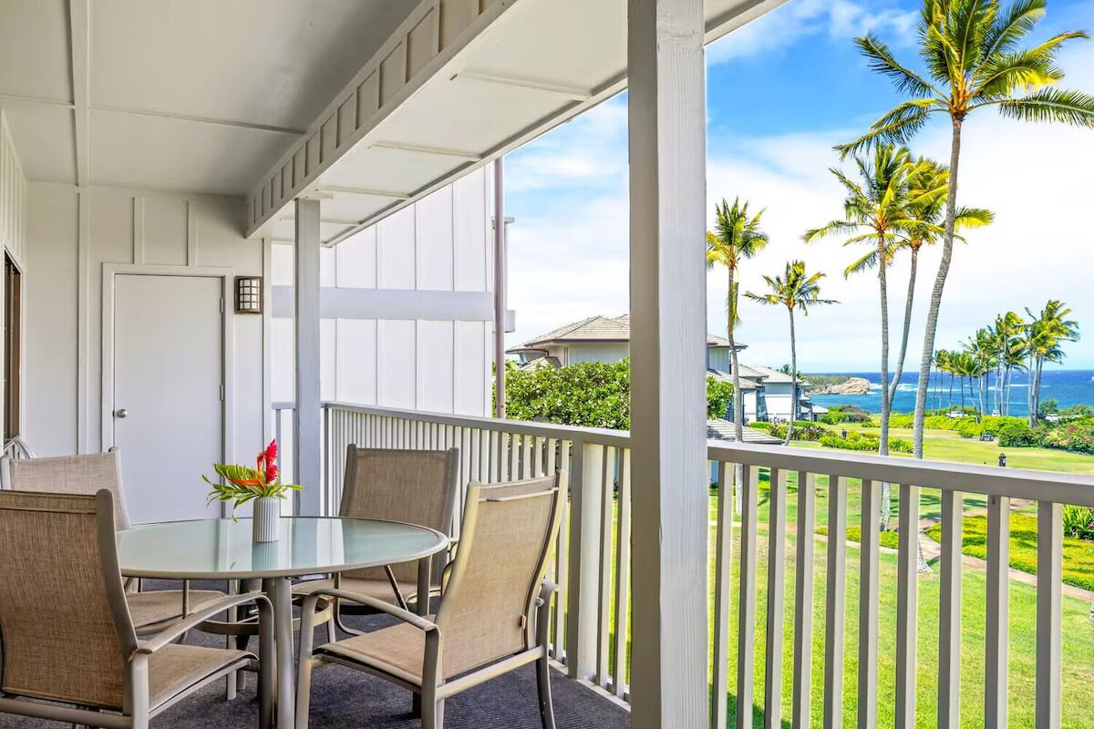 A balcony with a glass table and chairs, overlooking a well-maintained lawn and palm trees, with a view of the ocean and a building in the background.