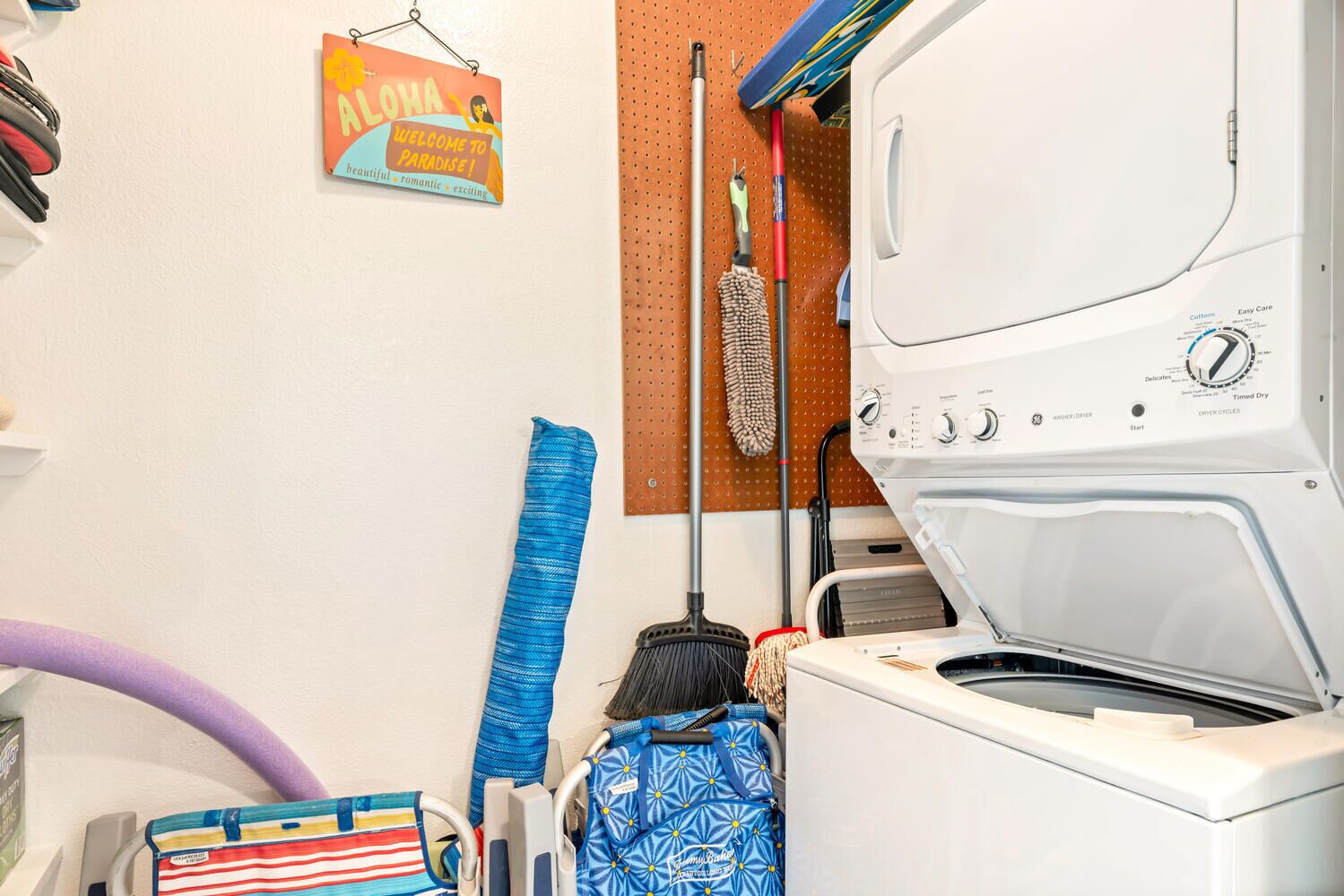 A laundry room with a washing machine, a mop, a broom, and a sign that says "ALOHA Welcome to Paradise!"