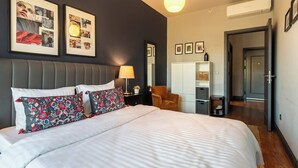Une chambre d’hôtel avec un grand lit, une table de chevet avec une lampe, une chaise et une armoire. Des tableaux encadrés ornent le mur et un appareil de climatisation est fixé au plafond.