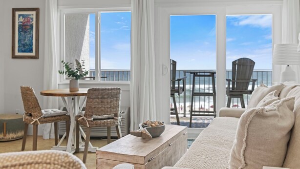 Un salón luminoso y espacioso con vista a la playa, que cuenta con muebles de mimbre, una mesa de centro de madera y un sofá beige.