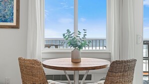 Una mesa redonda de madera con un jarrón blanco y una planta verde, acompañada por dos sillas de mimbre, en una habitación con un ventanal grande que da a un balcón.