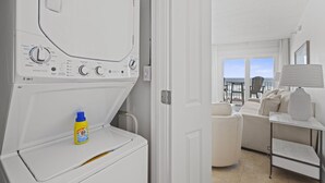 Un moderno lavadero blanco con lavadora y secadora de carga frontal, una botella de detergente y un balcón con vistas al mar.