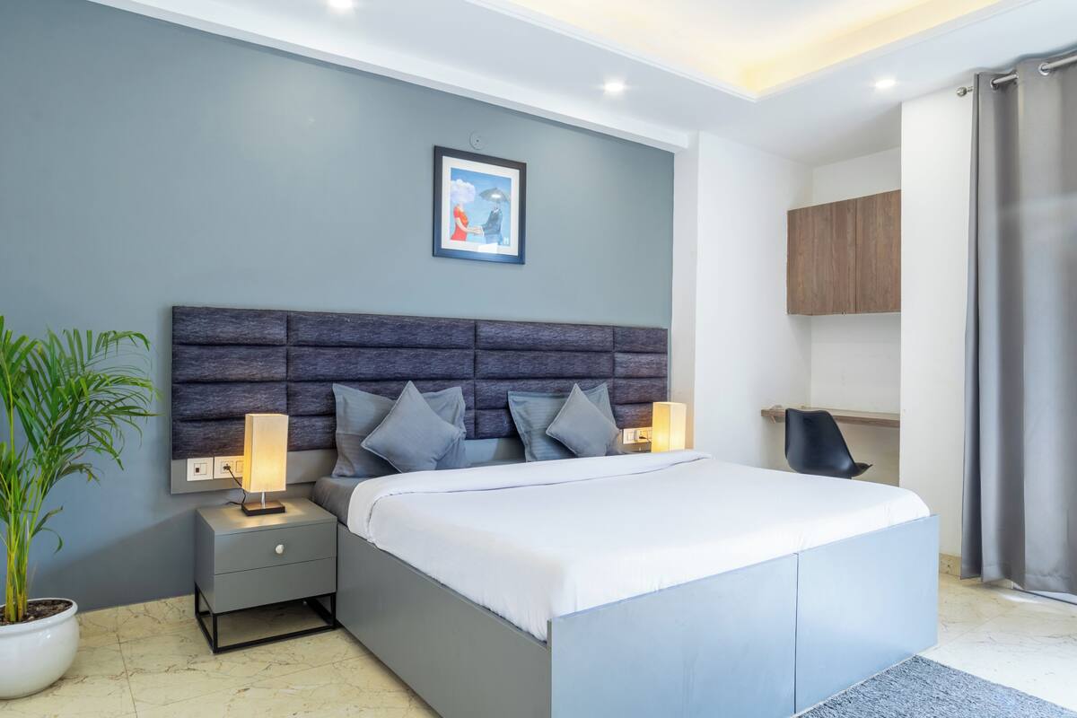 A modern bedroom with a large bed, bedside lamps, a nightstand, a potted plant, and a framed picture on the wall.