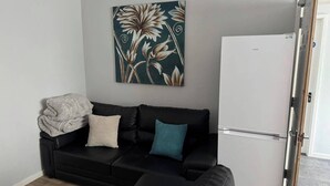 A living room with a black leather sofa, a white refrigerator, and a floral artwork on the wall. - 4 Bed Apartment in Warwick (Warwick)