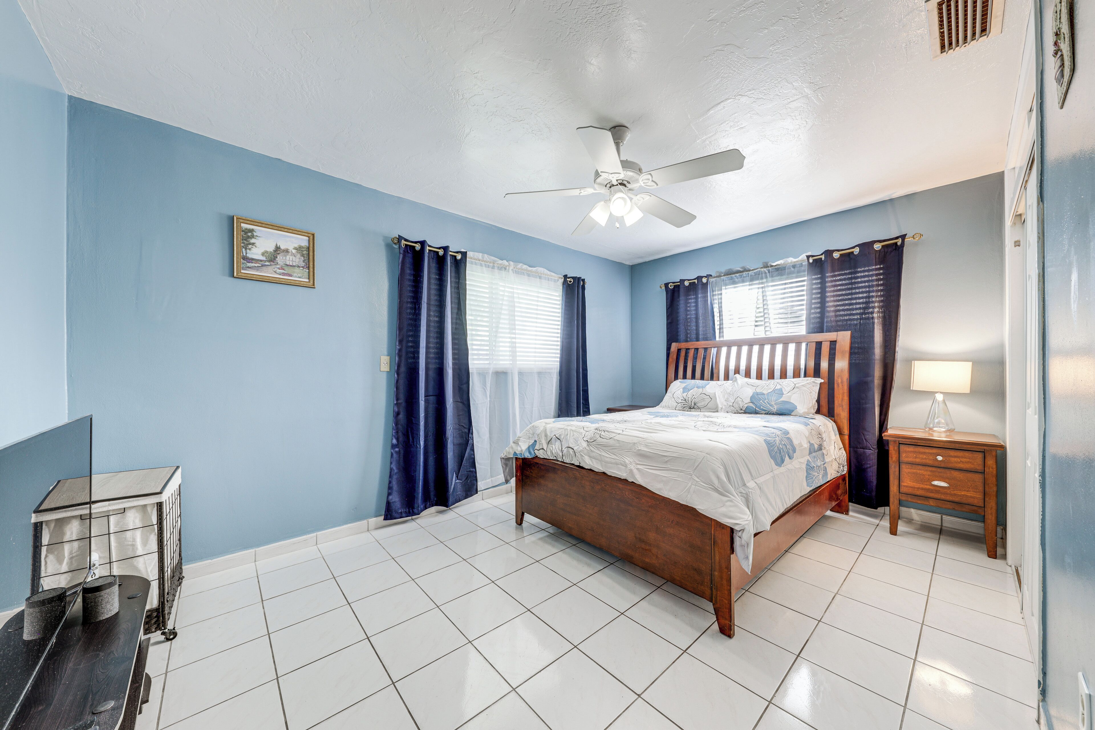 A bedroom with a wooden bed, a ceiling fan, a nightstand with a lamp, and a small table with a chair.