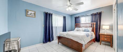 A bedroom with a wooden bed, a ceiling fan, a nightstand with a lamp, and a small table with a chair.