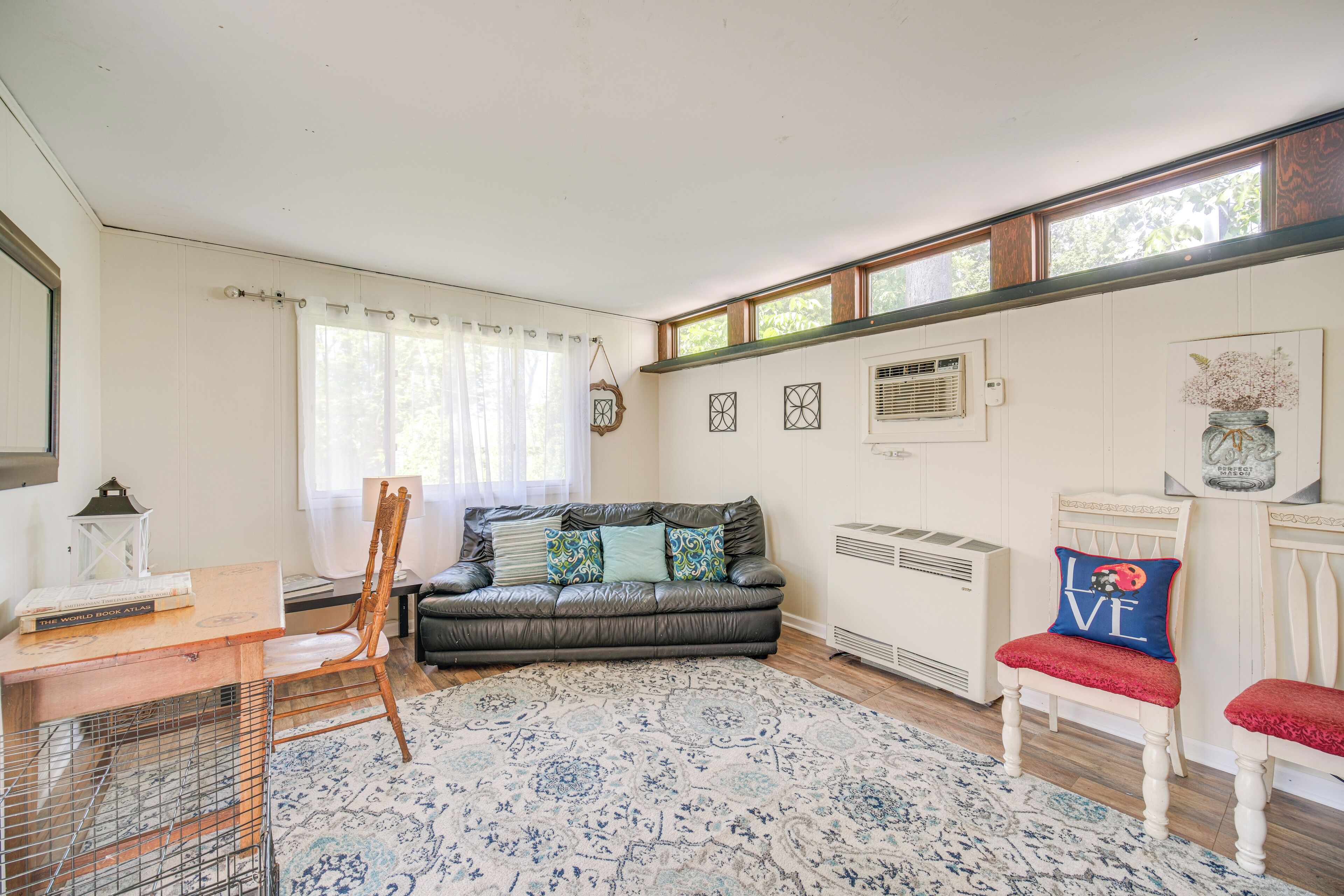 A room with a wooden desk, a leather sofa, a patterned rug, a white chair, and a large window with curtains.