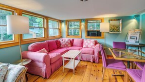 A cozy room with a pink sectional sofa, a small white table, a glass-top table, and a purple chair. There are large windows with wooden frames, a ceiling light, and a wall-mounted heater. - 13 Mi to Acadia: Artistic Retreat in Nature (Trenton)