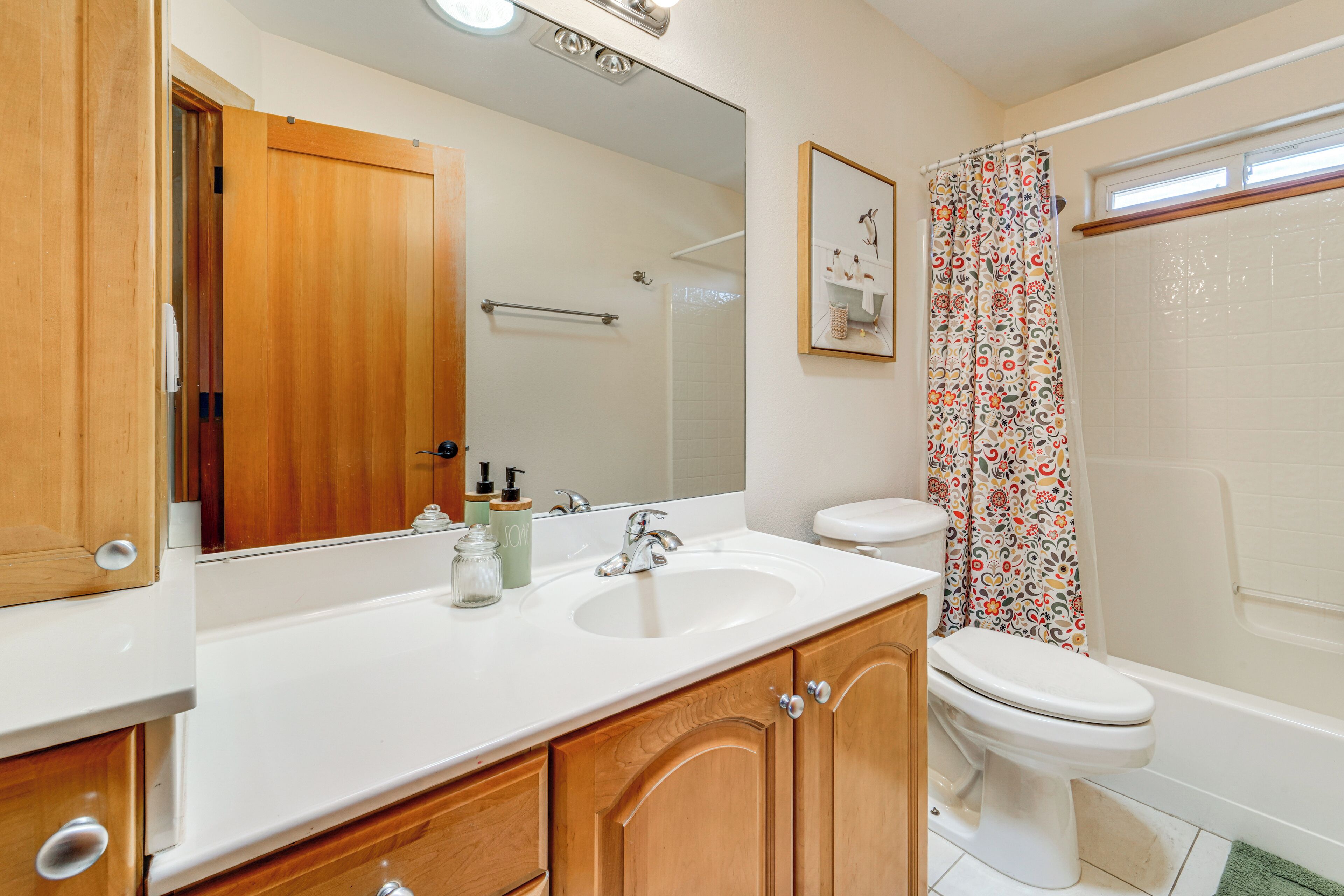 A bathroom with wooden cabinets, a white sink, a toilet, a shower with a floral curtain, and a mirror above the sink.