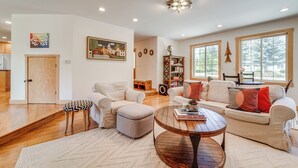 A spacious living room with a sofa, ottoman, and coffee table, a bookshelf, and large windows.