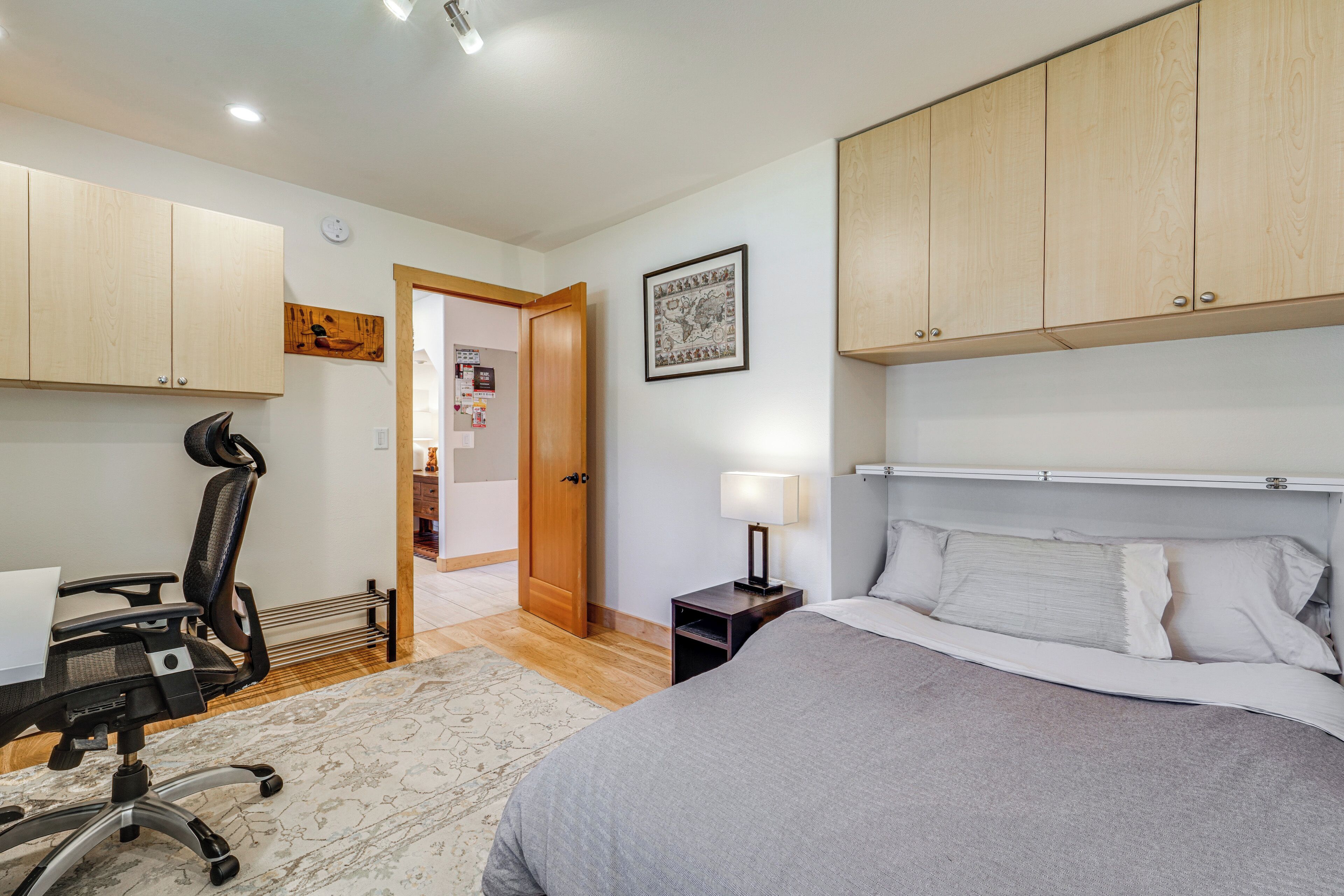 Un dormitorio con cama, escritorio con silla, armarios de madera, mesita de noche con lámpara y puerta que da a otra habitación.