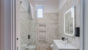 Un baño moderno con bidé, inodoro y lavamanos, paredes de mármol y un espejo grande.