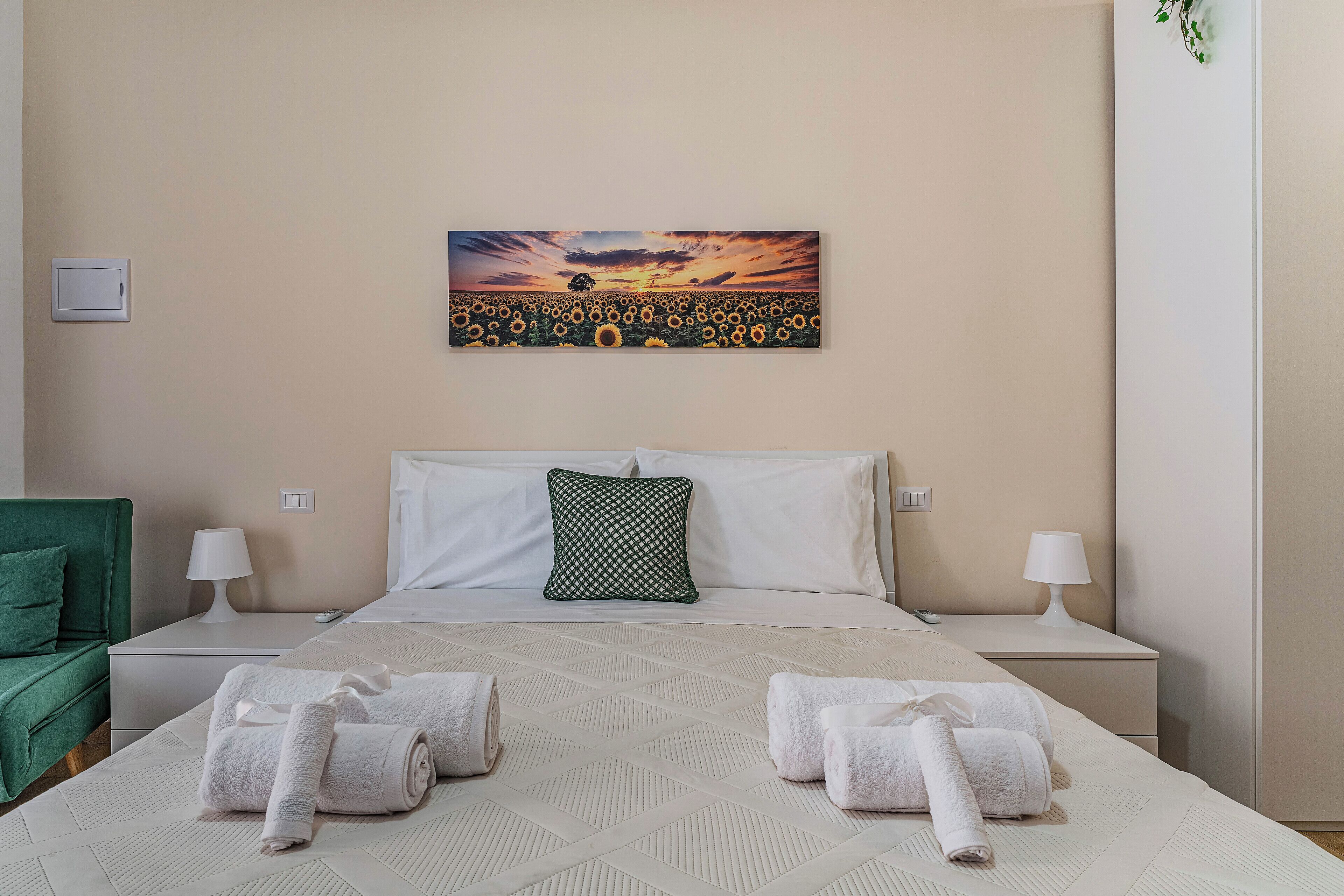 A hotel room with a bed, two nightstands, a green armchair, and a painting of a sunset over a field of sunflowers.