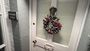 Una puerta blanca con un panel de vidrio esmerilado, un timbre de latón y un adorno con piñas y bayas rojas. Se ve un letrero que dice "Dunnmalt Cottage" en la pared a la izquierda.