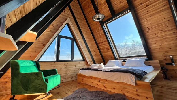 A cozy attic bedroom with a sloped ceiling, wooden beams, a bed, a green chair, and large windows. - Ordu Bungalov Loın Du Monde (Persembe)