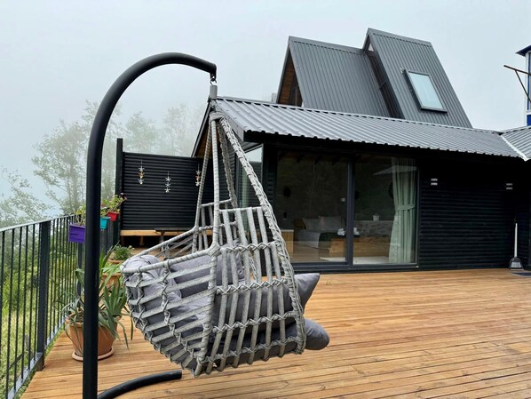 Une maison moderne dotée d’une terrasse en bois, d’un fauteuil suspendu et offrant une vue sur les environs.