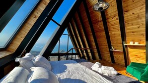 A bedroom with a sloped ceiling, wooden walls, a bed with white linens, and a view of the landscape through large windows. - Ordu Bungalov Loın Du Monde (Persembe)