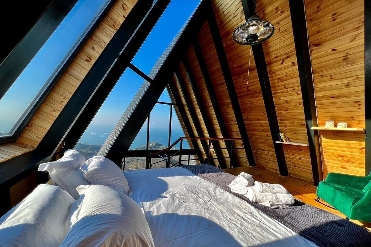 Ein Schlafzimmer mit schrägverdachter Decke, Holzwänden, einem Bett mit weißer Bettwäsche und Blick auf die Landschaft durch große Fenster.