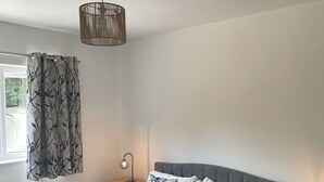 A neatly made bed with a grey headboard, a bedside table with a lamp, and a window with a curtain. - Hythe Hideaway - Home Away From Home (Hythe)