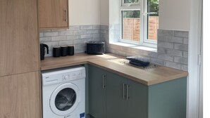 A compact kitchen with a washing machine, wooden cabinets, a window, and a hanging light fixture. - Hythe Hideaway - Home Away From Home (Hythe)