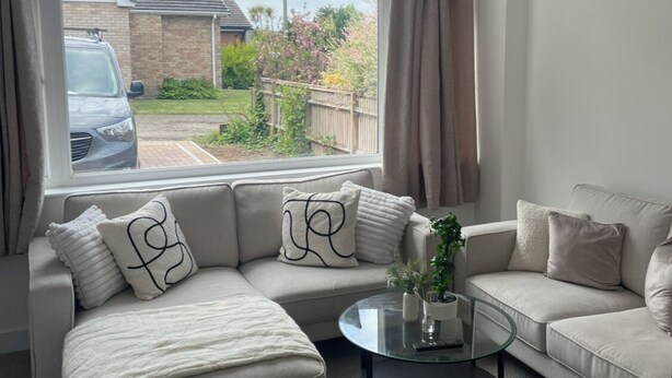 A living room with a beige sofa, a glass coffee table, and a view of a residential area through large windows. - Hythe Hideaway - Home Away From Home (Hythe)