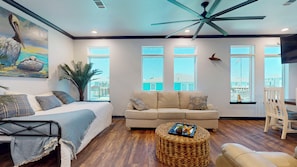 A spacious living room with a bed, sofa, and ottoman, large windows, and a ceiling fan.