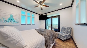 A bedroom with a bed, a chair, a ceiling fan, a TV, and a balcony with a view.