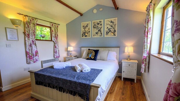 A bedroom with a bed, two bedside tables, a window with curtains, and wall-mounted artwork. - Bluebells by STAE-Homes (Bristol)