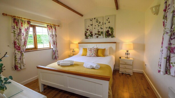 A bedroom with a wooden bed, white headboard, floral curtains, a bedside table with a lamp, and a window with a view of greenery. - Lavender by STAE-Homes (Bristol)