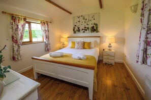 A bedroom with a wooden bed, white headboard, floral curtains, a bedside table with a lamp, and a window with a view of greenery.