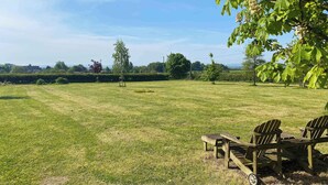 A grassy field with a wooden bench and chairs, a picnic table, and a tree with green leaves. - Rose by STAE-Homes Su (Bristol)