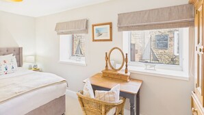 A bedroom with a bed, a dresser with a mirror, a wicker chair, and two windows with curtains. - Host & Stay | St Cuthbert's Cottage (Seahouses)