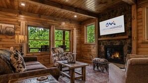 A cozy log cabin interior with a fireplace, wooden furniture, and large windows offering a view of the forest.