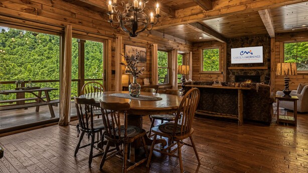 A wooden cabin interior with a dining table, chairs, a chandelier, a fireplace, and large windows overlooking a forest.