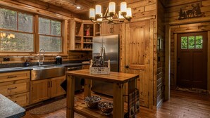 A wooden cabin kitchen with a stainless steel refrigerator, wooden cabinets, a wooden dining table, and a window overlooking the forest.
