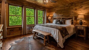 A wooden cabin bedroom with a bed, bench, and two windows offering a view of greenery.