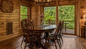 A wooden interior with a round dining table, four chairs, a chandelier, and a view of greenery outside.