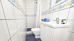 A bathroom with white tiles, a blue toilet, a white sink with a mirror, and a small plant on the sink.