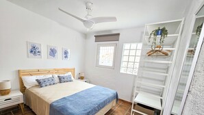 A bedroom with a bed, a nightstand, a bedside table, a ceiling fan, a window, and a shelving unit.