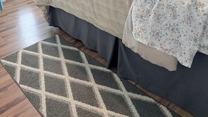 Una camera da letto con un letto, una TV fissata al muro, un comodino con un vaso e una cornice, e una finestra con tende.
