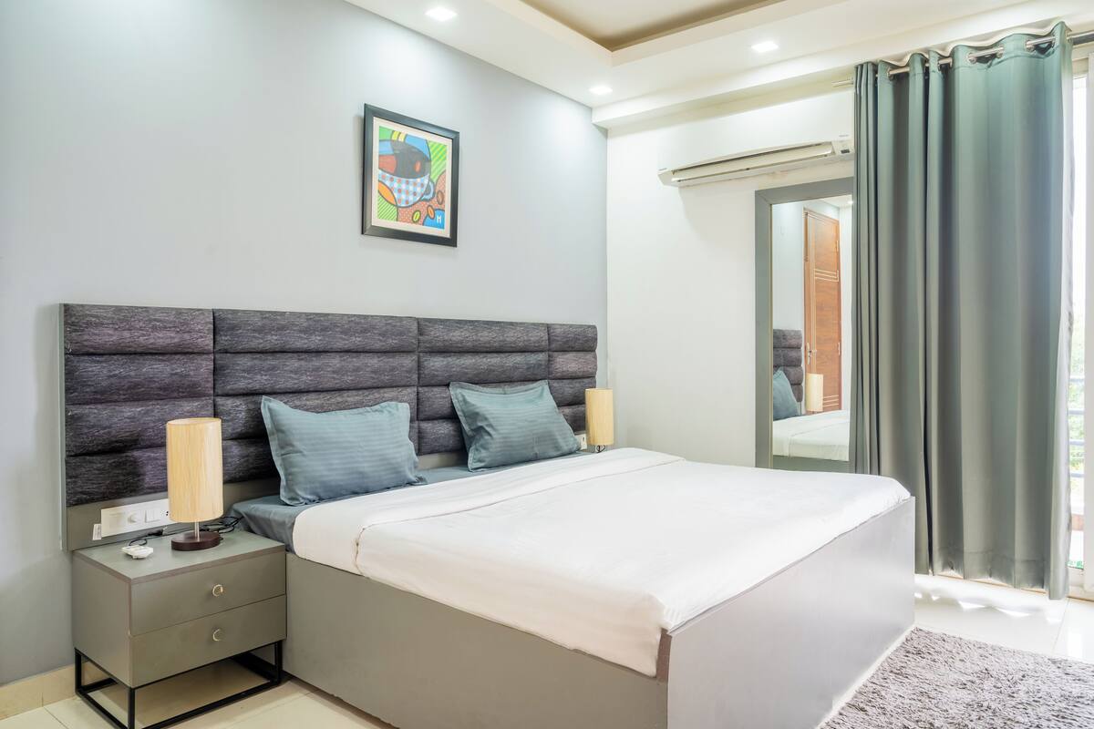 A modern bedroom with a grey headboard, two bedside tables, a bed with white bedding, a lamp, and a framed artwork on the wall.