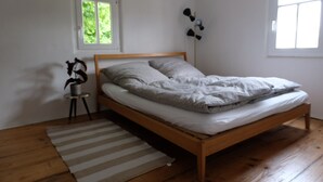 Un lit simple avec un cadre en bois, dans une pièce aux murs blancs et dotée d’une fenêtre.