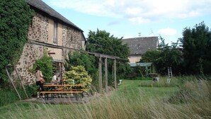 Un bâtiment rustique en pierre, agrémenté d’une pergola en bois et de tables de pique-nique, situé dans un espace verdoyant.