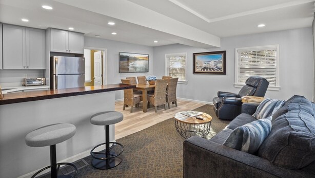 A modern kitchen with a central island, bar stools, a dining area with a table and chairs, a leather sofa, and a coffee table. - Unique West Harwich Vacation Compound w/ Pool RPAM111 (West Harwich)