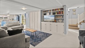 A spacious living room with a grey sofa, a coffee table, and a bookshelf filled with books and decorative items. - Unique West Harwich Vacation Compound w/ Pool RPAM111 (West Harwich)